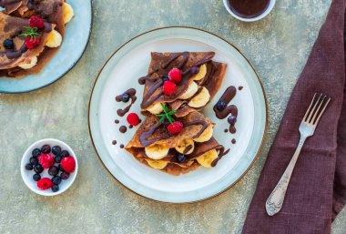 Muz, ahududu ve yaban mersiniyle doldurulmuş ince çikolatalı krepler, açık beton arka planda gri bir seramik tabağa çikolata sosu dökülüyor. Krep tarifleri. Krep günü.