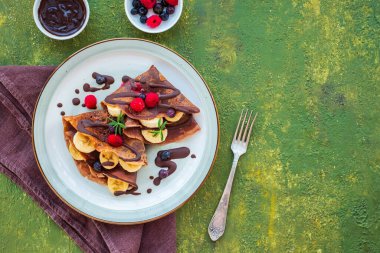 Muz, ahududu ve yaban mersiniyle doldurulmuş, çikolata soslu, yeşil beton arka planda gri bir seramik tabağa serpiştirilmiş ince çikolatalı krepler. Krep tarifleri. Krep günü.