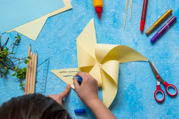 Step-by-step making of a paper weather vane by a child on a blue concrete background. Children\'s creativity, divas, crafts. Paper crafts