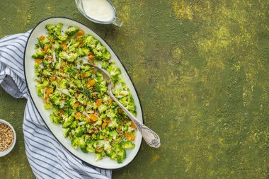 Sağlıklı bir çiğ brokoli salatası, soğan, kuru üzüm ve yeşil beton arka planda beyaz oval tabakta yoğurt soslu ayçekirdeği. Salata tarifleri, vejetaryen yemekleri. Çiğ yemek.