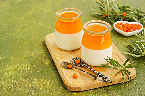 Десерт, сливочная панна котта с облепиховым желе в винтажных стеклянных банках на деревянной доске или поднос на оливково-зеленом бетонном фоне. Рецепты облепихи. Итальянские десерты.