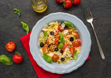 Siyah beton arka planda gri bir tabakta makarna, domates, zeytin ve parmesan salatası. Makarna salatası tarifleri. İtalyan yemeği..