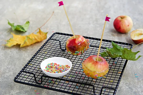 Dessert, caramelized candy apples in transparent caramel, sprinkled with sugar decor, on a black grate on a gray concrete background. Apples recipes. Party food. Children\'s holiday.