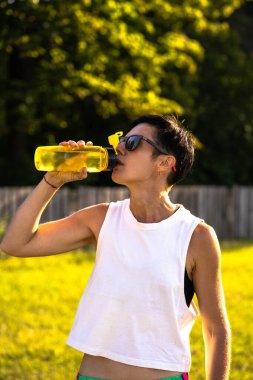 Genç, güzel bir kadın antrenmandan sonra parkta plastik şişeden su içiyor, kısa siyah saç, güneş gözlüğü ve şort, yazın açık havada spor, spor kıyafetlerinde ince formalı kız, spor yaparken kadın, dikey çekim, fotokopi alanı.