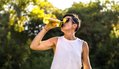 Genç, güzel bir kadın antrenmandan sonra parktaki plastik şişeden su içiyor. Kısa siyah saç, güneş gözlüğü ve şort. Yazın açık havada spor yapıyor.