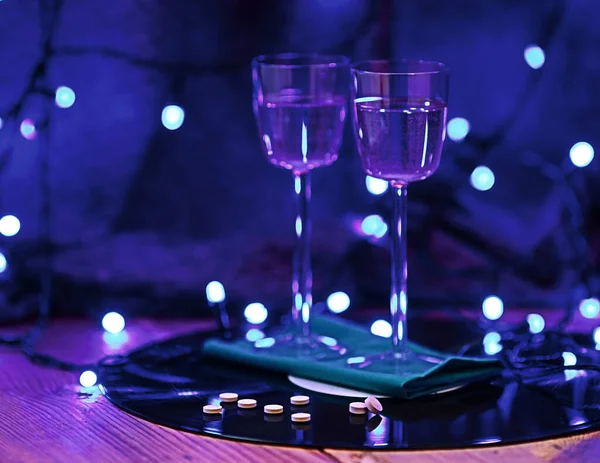 selective focus photo of pills against the background of two glasses of champagne and a vinyl record, surrounded by bright club lighting