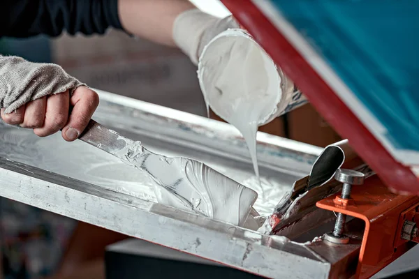 Erkek ellerinin spatulası ve beyaz boya kutusuyla seçici odak fotoğrafı. serigrafi üretimi. Bir tasarım stüdyosunda ipek ekran yöntemiyle tişörtlere resimler basmak
