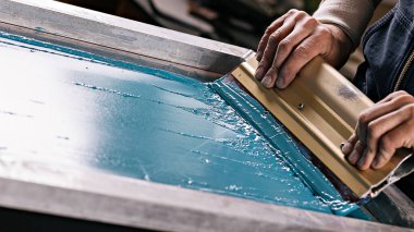 Seçici odaklı erkek eli ve cam sileceği fotoğrafı. serigrafi üretimi. Bir tasarım stüdyosunda ipek ekran yöntemiyle tişörtlere resimler basmak