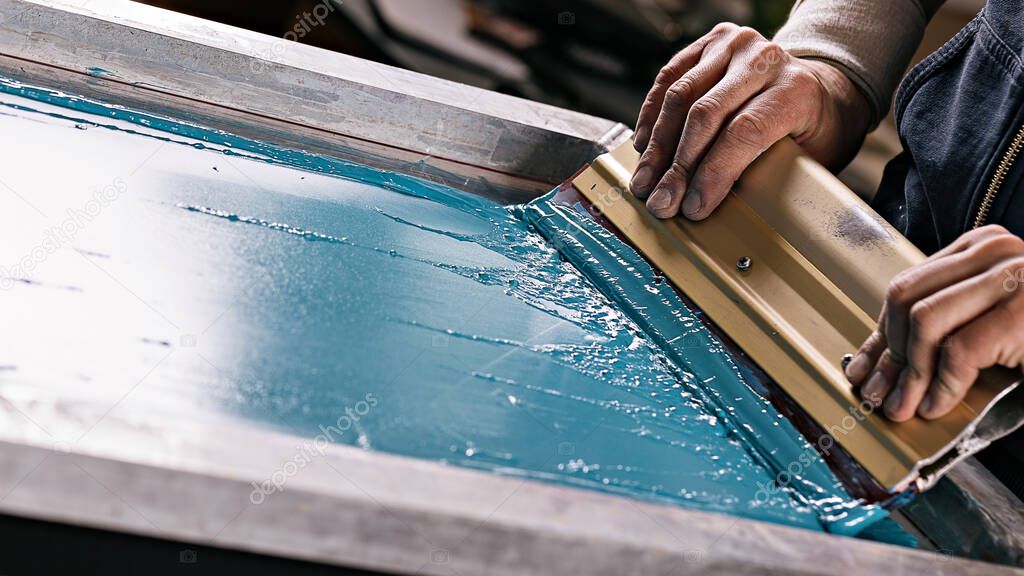 selective focus photo of male hands with squeegee. serigraphy production. printing images on t-shirts by silkscreen method in a design studio