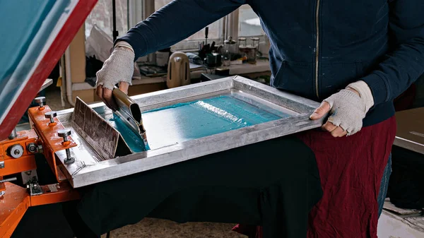 Seçici odaklı erkek eli ve cam sileceği fotoğrafı. serigrafi üretimi. Bir tasarım stüdyosunda ipek ekran yöntemiyle tişörtlere resimler basmak
