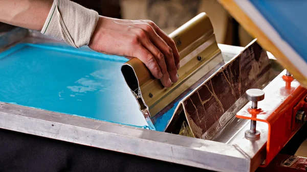 Seçici odak fotoğrafı, erkek eli ve cam sileceği. serigrafi üretimi. Bir tasarım stüdyosunda ipek ekran yöntemiyle giysiler üzerine resimler yazdırmak