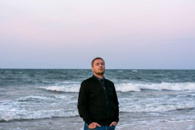 Genç beyaz adam akşamları sahilde duruyor. Gün batımında beyaz köpüklü büyük dalgalar. Seçici odak.