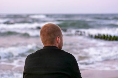 Genç adam akşamları gün batımında sırtı deniz kıyısında oturur. Seçici odak, bulanık.