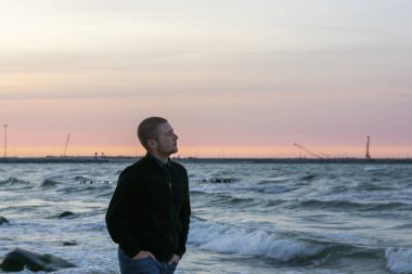 Genç beyaz adam gün batımında deniz kıyısında yürüyor ve beyaz köpükle dalgalara bakıyor. Seçici odak, bulanık