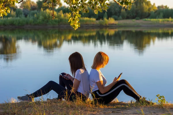 Duas Jovens Mulheres Sentam Margem Rio Olham Para Smartphones Concepção — Fotografia de Stock