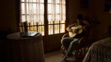 Evde dizüstü bilgisayarla gitar çalmayı öğrenmek. Mutlu genç adam evdeki kanepede otururken gitar çalıyor..