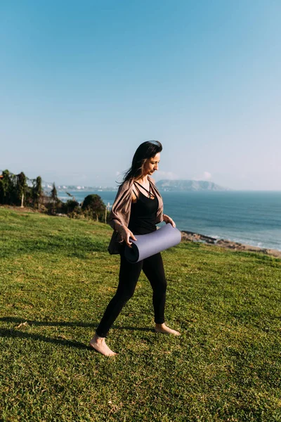 Kadın yoga minderini denizin önündeki çimlere koymaya hazırlanıyor. Boşluğu kopyala