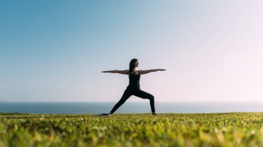 Kollarını gökyüzü ve denizin arka planında açmış yoga yapan bir kadın. Boşluğu kopyala