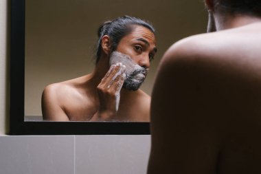 Saç kurdelesi takan genç Latin adam banyoya tıraş köpüğü koyuyor.