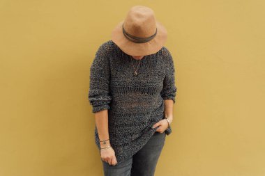 Şapkalı bir kadın bir eli cebinde, diğeri de sarı hardal duvarına yaslanmış kıyafetlerini çekiyor. Şehir modası konsepti. Boşluğu kopyala