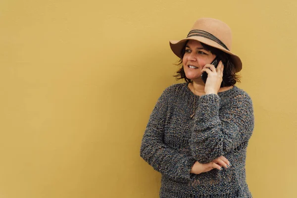 Bir kadın telefonuyla konuşurken gülümsüyor, şapka takıyor ve sarı bir duvara yaslanıyor. Şehir görüntüsü