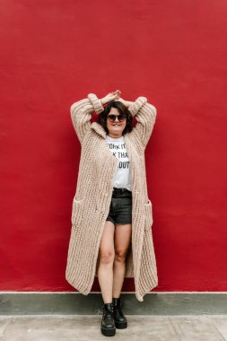 Uzun süveterli, siyah gözlüklü, şortlu, kolları kırmızı duvara dayanmış siyah ayakkabılı bir kadın. Dikey resim