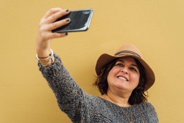 Cep telefonuyla fotoğraf çeken bir kadın. Kazaklı kadın sarı duvara yaslanıyor.