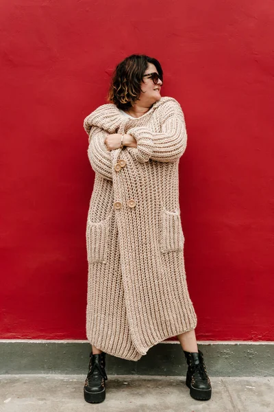 Woman covers her body with a long skin-colored sweater, wears sunglasses, black boots and looks sideways against a red background