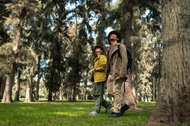 İki kişi ormanda yürüyor, biri sarı ceketli, diğeri kahverengi ceketli.