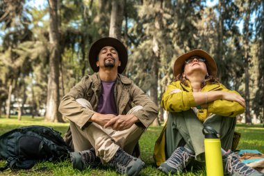 Birkaç turist ormanda oturmuş, gözleri kapalı mekanın tadını çıkarıyorlar. Şapka, sırt çantası takıyorlar ve su şişesi tutuyorlar.