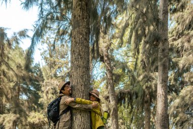 İki kişi ormanda bir ağaca sarılıyor. Adam sırt çantası takıyor ve kadın sarı bir ceket giyiyor. Anın ve doğanın güzelliğinin tadını çıkarıyor gibiler.