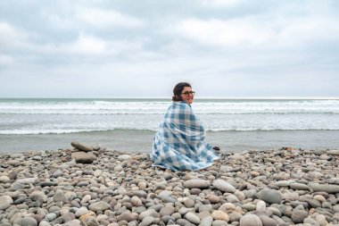 Bir kadın kumsalda oturuyor ve battaniyesi etrafında. Gökyüzü bulutlu ve arkada okyanus var.