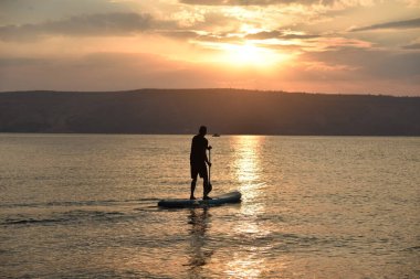 Güneş doğarken büyük bir gölde SUP üzerinde yelken açan bir adam. Ayağa kalk ve kürek çek - doğada aktif eğlence.. 