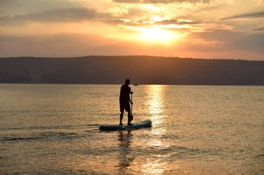 Güneş doğarken büyük bir gölde SUP üzerinde yelken açan bir adam. Ayağa kalk ve kürek çek - doğada aktif eğlence.. 