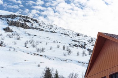 Sığ karla kaplı bir dağ. Kardan süzülen çalılar. Bulutlu mavi gökyüzü. Oduncu kulübesi. Ahşap kulübe