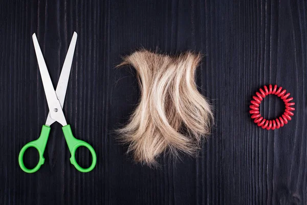 Blond lock of hair, scissors, scrunchy black wooden background closeup, cut off blonde hair curl on dark wood, shears, spiral hair band barrette, hairstyle accessories, haircut, barber, hairdo concept