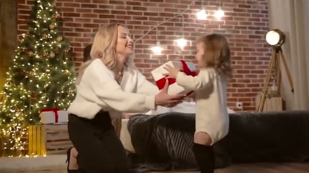 A cute daughter girl runs up to her mother and gives a Christmas present. Mom and little child are having fun indoors near a tree. Loving family with gifts in the room. Merry Christmas and Happy — Stock Video