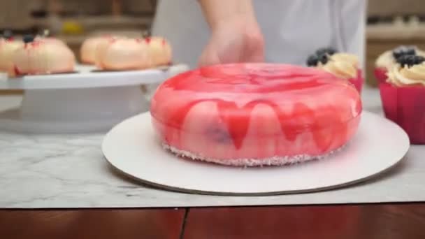 Mujer pastelera en su cocina pone un pastel en esmalte rojo en la mesa con postres cocidos, sonrisas. Happy chef pone pastel terminado en la mesa con cupcakes, para las vacaciones — Vídeo de stock
