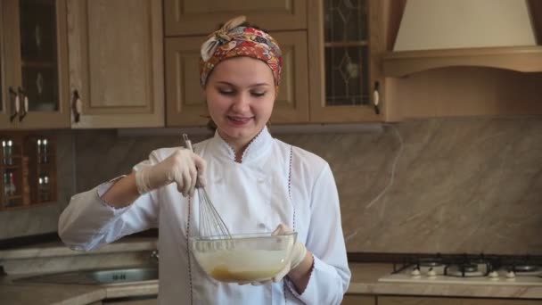 Η γυναίκα χαμογελά και ζυμώνει ζύμη με σύρμα σε χορταστικό μπολ. Στη μαγειρική, μια γυναίκα αναμιγνύει τα συστατικά σε γυάλινο μπολ με ένα σύρμα — Αρχείο Βίντεο