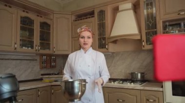 Mutfakta yemek pişiren kadın hamur makinesindeki malzemeleri karıştırıyor, yemek tarifini anlatıyor ve akıllı telefonuyla yemek pişirme blogu için video çekiyor.