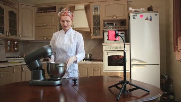 Chef de pastelaria mulher na cozinha diz um reset para um blog de culinária. Mulher diz receita, amassa a massa de farinha com misturador e dispara no smartphone do blog vídeo — Vídeo de Stock