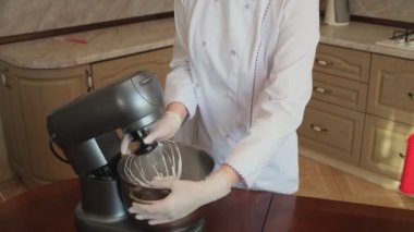 Mutfaktaki bir kadın pasta şefi yemek blogu için telefonuna video çekiyor, bir elektrikli miksere krema karıştırıyor, süzüyor ve un ekliyor. Kadın blogcu krema pişirmenin yolunu söyler