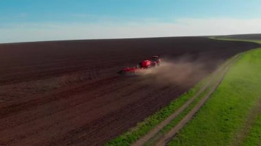 Ekilebilir, kırmızı traktör toprağı kazar ve ilerler, arkasından bir toz sütunu yükselir, güneşli bir gün, parıltı, tarım alanı, insansız hava aracının manzarası