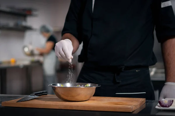 Aşçı elleri eldivenle yemek pişirir. Mutfakta Yunan salatasına tuz ekler.