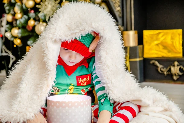 Милый маленький мальчик, одетый эльфом, открывает рождественский подарок, прячась за пушистым одеялом в украшенном рождественском доме.. — стоковое фото