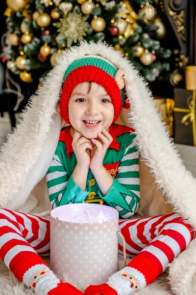 A cute little boy dressed as an elf opens a Christmas present while hiding behind a fluffy blanket in a decorated Christmas house. — Stock Photo, Image