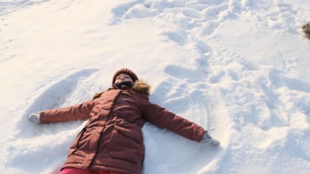 O fetiță sănătoasă face un înger de zăpadă în parc pe zăpadă. Copilărie fericită, plimbări de iarnă în pădure . — Videoclip de stoc
