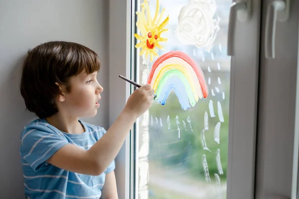 Un niño pequeño dibuja un arco iris en una ventana durante una pandemia de coronavirus. — Foto de Stock