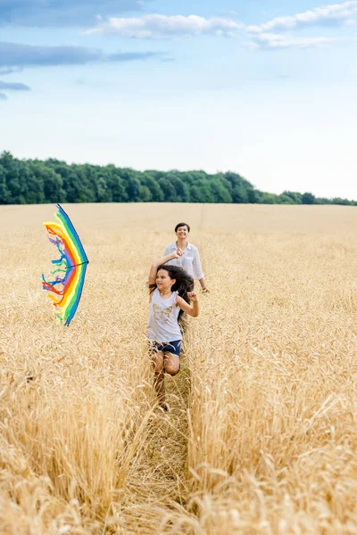 Mãe e filha correm em um campo de trigo com um papagaio no verão. Fim de semana bem planeado e activo. Infância feliz. — Fotografia de Stock