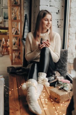 Genç ve güzel bir kadın bulutlu bir sabahta elinde bir fincan kahveyle pencere eşiğinde oturuyor. Bulutlu bir sabahta sıcak kahve.